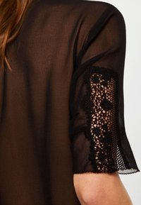 Vêtement noir transparent à manches courtes avec des détails en dentelle noire complexe aux poignets. Texture douce et design fluide.