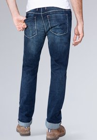 Jeans in denim blu scuro con vestibilità dritta, caratterizzati da cuciture chiare, orli arrotolati e due tasche posteriori. Indossati con scarpe in suede marrone.