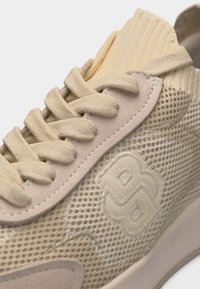 Gros plan sur une chaussure de sport en maille beige avec col rembourré, lacets épais, superpositions en daim et logo en relief sur le côté.