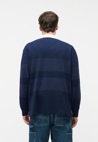 Άνδρας που φοράει μπλε ναυτικό μακρυμάνικο πουκάμισο ράγκμπι με δίχρωμες οριζόντιες ρίγες και μπλε τζιν, στέκεται γυρισμένος με την πλάτη προς ένα απλό φόντο.