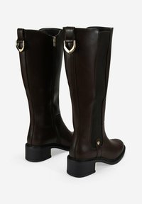 Botas de cuero marrón hasta la rodilla con un panel trasero texturizado, detalles en hardware dorado y un tacón de bloque modesto. Detalle de cremallera en el costado.