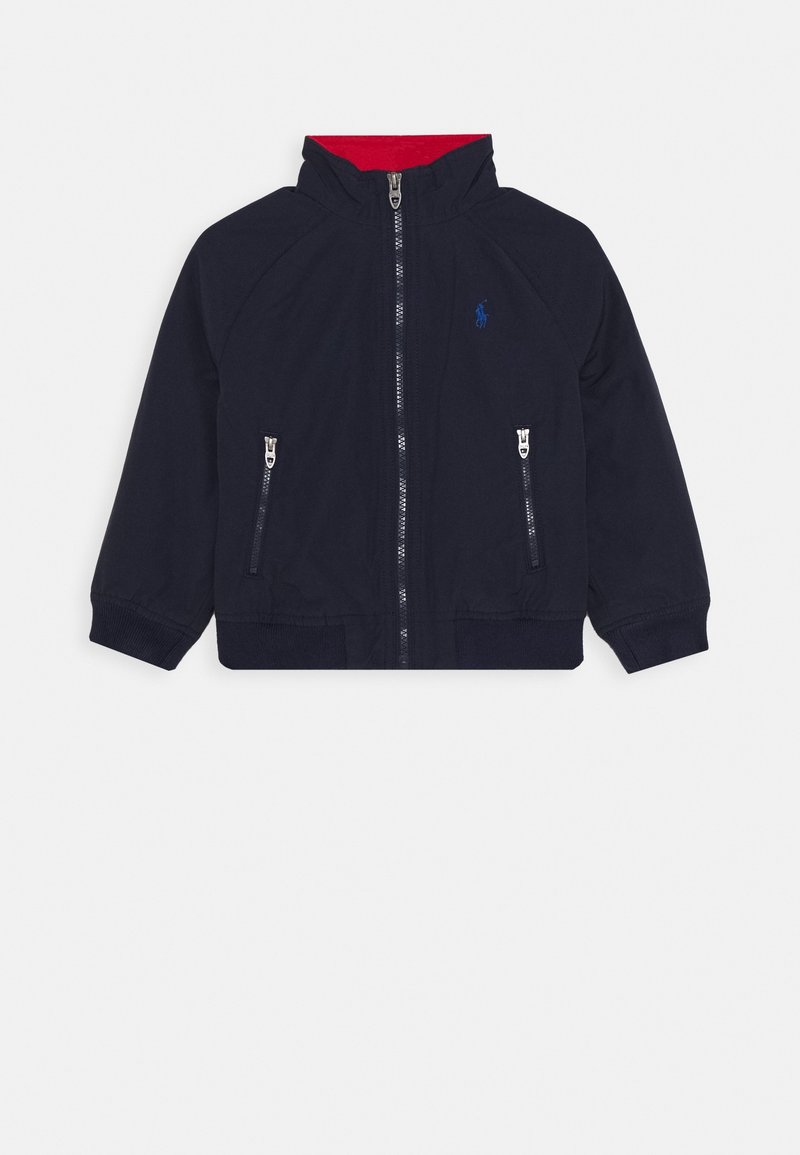 Marineblauwe kinderjas met rode kraagvoering, een rits aan de voorkant, twee ritszakken en een klein blauw geborduurd logo op de borst.