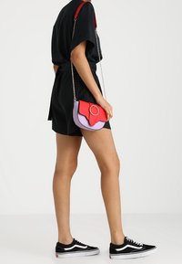 Barevná křížová taška s červeným a levandulovým designem, zakřivenými okraji a kulatým kovovým detailem, připevněná na řetízkovém popruhu.