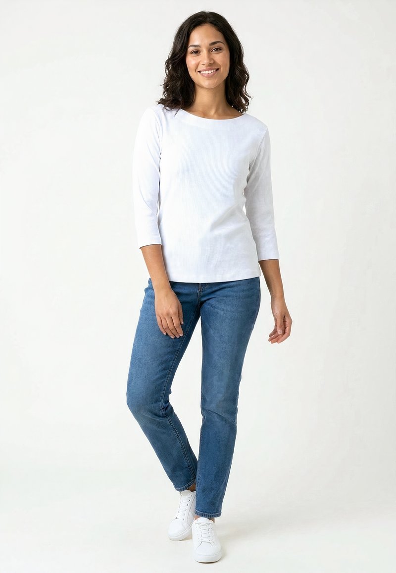 Femme souriante aux cheveux foncés portant un haut blanc à manches longues, un jean bleu et des baskets blanches, debout devant un fond clair uni.