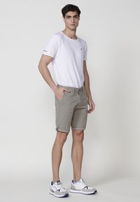 Tricou ușor din bumbac alb, cu decolteu rotund, asortat cu pantaloni scurți de culoare verde măsliniu, cu margini rulate și buzunare. Poartă adidași albi și gri.