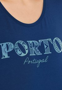 Tmavě modré tričko s kruhovým výstřihem, na kterém je nápis "PORTO" v dekorativních světle modrých písmenech a "Portugal" ve zdobeném písmu.