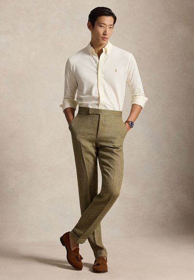 Mees seisab käed taskutes, kannab kreemikat nööpidega särki, oliivirohelisi kõrge vöökohaga pükse, pruune tuttidega loafereid ja käekella.