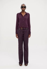 Camicia a maniche lunghe viola scuro con bottoni, abbinata a pantaloni alti dello stesso colore e una cintura. Gli accessori includono occhiali da sole scuri e sandali con tacco.