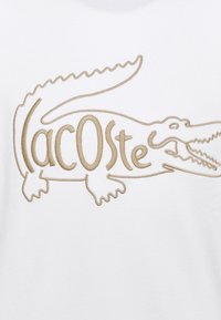 Tissu blanc avec un logo crocodile brodé en beige, accompagné du texte stylisé « Lacoste » intégré au design.