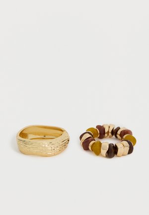 Guldtekstureret chunky ring ved siden af et elastikarmbånd med skiftevis guldfarvede, brune og sennepsfarvede stenlignende perler på hvid baggrund.