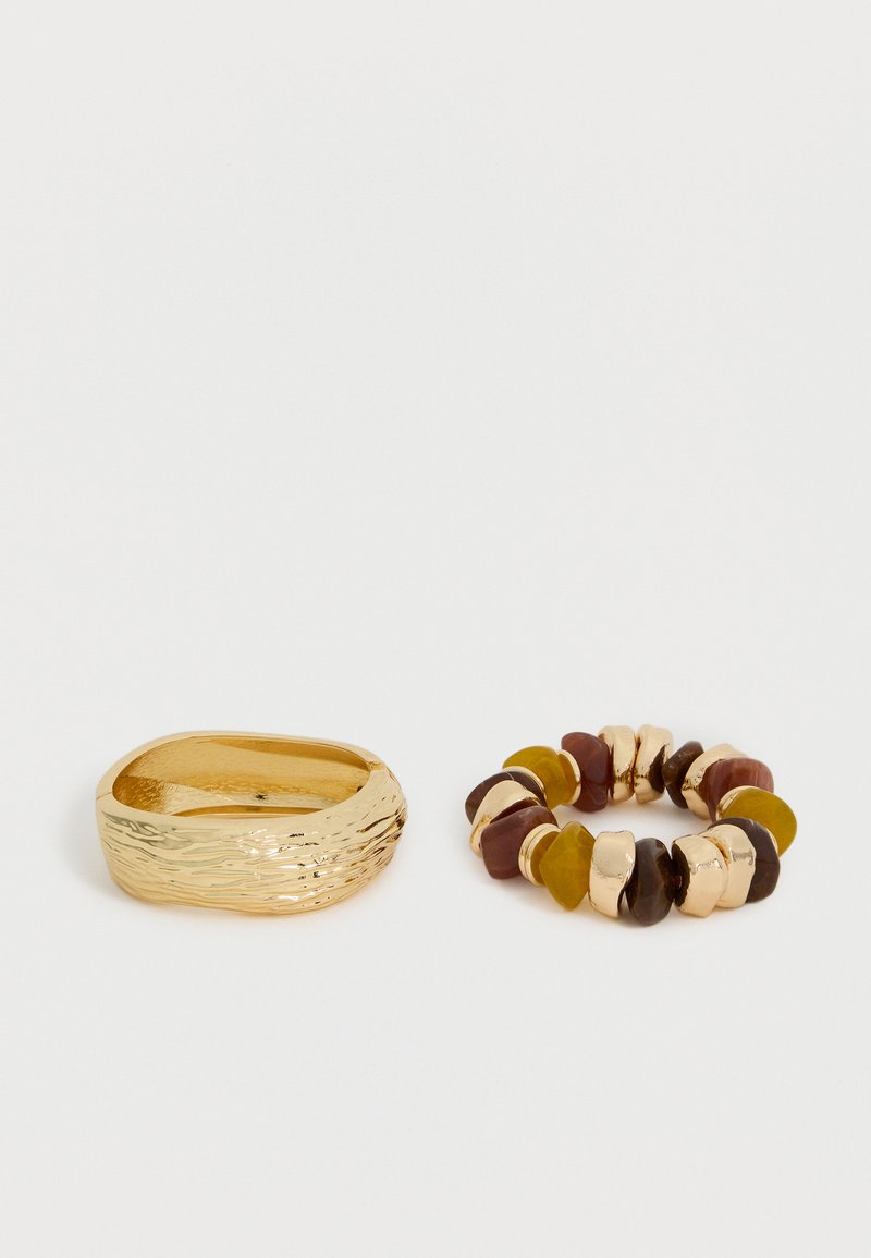 Anneau épais texturé en or à côté d'un bracelet élastique avec des perles alternées ressemblant à des pierres de couleur or, marron et moutarde sur un fond blanc.