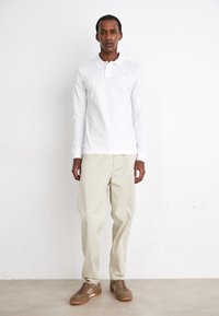 Cămașă polo albă cu mânecă lungă, pantaloni bej largi și adidași maro. Design minimalist cu forme simple și țesături moi.
