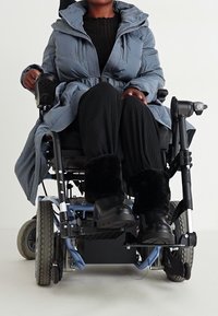 Electric wheelchair with blue frame, black wheels; user wearing a blue puffy coat, black pants, and black fur-lined boots. Clear, neutral background.