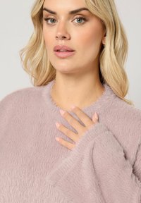 Femme blonde avec un maquillage léger portant un pull en lilas duveteux, touchant sa poitrine avec la main montrant des ongles manucurés rose pastel, arrière-plan neutre.