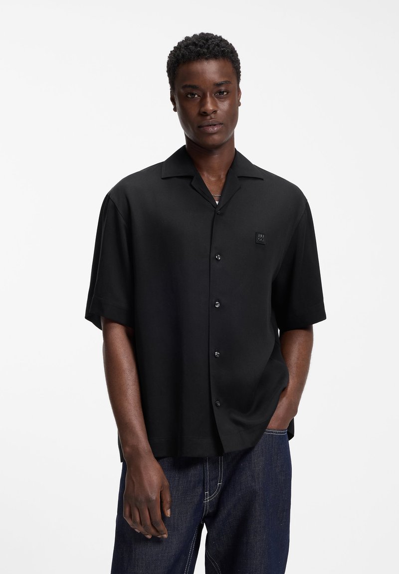 Νέος άνδρας που φοράει μαύρο πουκάμισο με κοντό μανίκι και κουμπιά και σκούρο μπλε τζιν παντελόνι, στέκεται με το ένα χέρι στην τσέπη μπροστά σε λευκό φόντο.