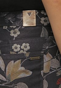 Vaqueros de mezclilla con estampado floral, pequeña etiqueta metálica "GUESS" en el bolsillo y parche de cuero beige con la marca "GUESS Los Angeles" en la cintura.