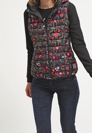 Kvinde iført sort langærmet skjorte og sort quiltet vest med røde, pink og hvide blomsterprint, kombineret med mørkeblå jeans.