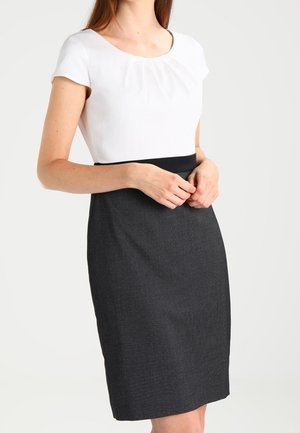 Kobieta ubrana w biały krótki rękaw wsunięty w ołówkową spódnicę z wysokim stanem, stoi z rękami złączonymi na wysokości talii.