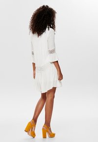 White dress with flared sleeves, lace accents, and pleats. Worn with yellow platform heels, showing a soft texture and flowy design.