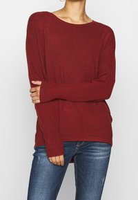 Pull bordeaux à manches longues en tissu tricoté texturé. Il présente un large décolleté et des poignets ajustés, associé à un jean bleu délavé et usé.