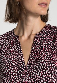 Femme aux cheveux bruns courts portant un blouse à motif noir, blanc et rouge avec un décolleté en V, devant un fond uni.
