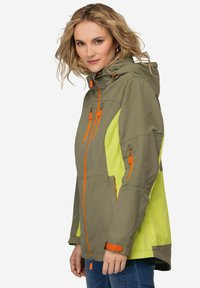 Chaqueta ligera y resistente al agua en verde olivo y amarillo neón, con cremalleras naranjas y puños y capucha ajustables. Múltiples bolsillos con cremallera.