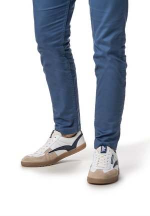 White and navy sneaker with suede accents and a beige rubber sole; paired with blue pants, showing a casual, sporty look.