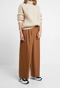Beige gerippter Pullover mit Hohem Kragen, kombiniert mit braunen weiten Hosen und schwarz-weißen Sneakers. Gemütliche Texturen mit lässigem Schnitt.