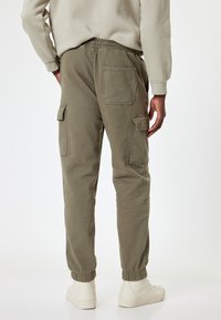 Mężczyzna w oliwkowych spodniach cargo z kieszeniami i elastycznymi mankietami, beżowej bluzie i białych trampkach, stojący plecami do aparatu.