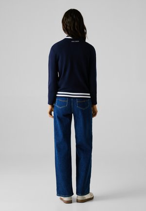 Pull marin avec des rayures blanches au bas et aux poignets, associé à un jean bleu ample et des chaussures de couleur claire, vu de l'arrière.