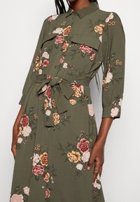 Olivgrünes Kleid mit floralem Muster in Pink, Gelb und Rot. Mit Knopfleiste, zwei Brusttaschen und einem Stoffgürtel in der Taille.