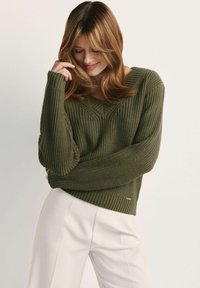 Sukienka w kolorze oliwkowym, dziana, z dekoltem w serek i prążkowanym wykończeniem. Luźny krój, długie rękawy i akcenty na dolnym wykończeniu. Zestawiona z białymi spodniami.