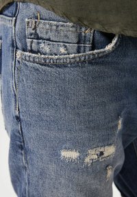 Vaqueros de mezclilla con un lavado azul medio, con parches desgastados, bordes deshilachados y diseño de cinco bolsillos. Detalles de costura prominentes visibles.