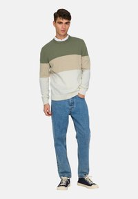 Pull en maille à manches longues avec rayures horizontales en vert olive, beige et blanc. Associé à un jean en denim bleu et des baskets bleu marine.