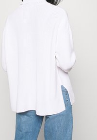 Weißer Strickpullover mit hohem Kragen, weiten Ärmeln, seitlichen Schlitzen und strukturiertem Muster, kombiniert mit hellblauen Jeans.