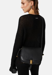 Borsa a tracolla nera in pelle con forma arrotondata, dotata di hardware in tonalità oro e una tracolla annodata, abbinata a un maglione in maglia nera con trama testurizzata.