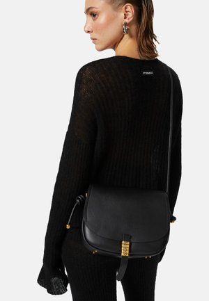 Borsa a tracolla nera in pelle con forma arrotondata, dotata di hardware in tonalità oro e una tracolla annodata, abbinata a un maglione in maglia nera con trama testurizzata.