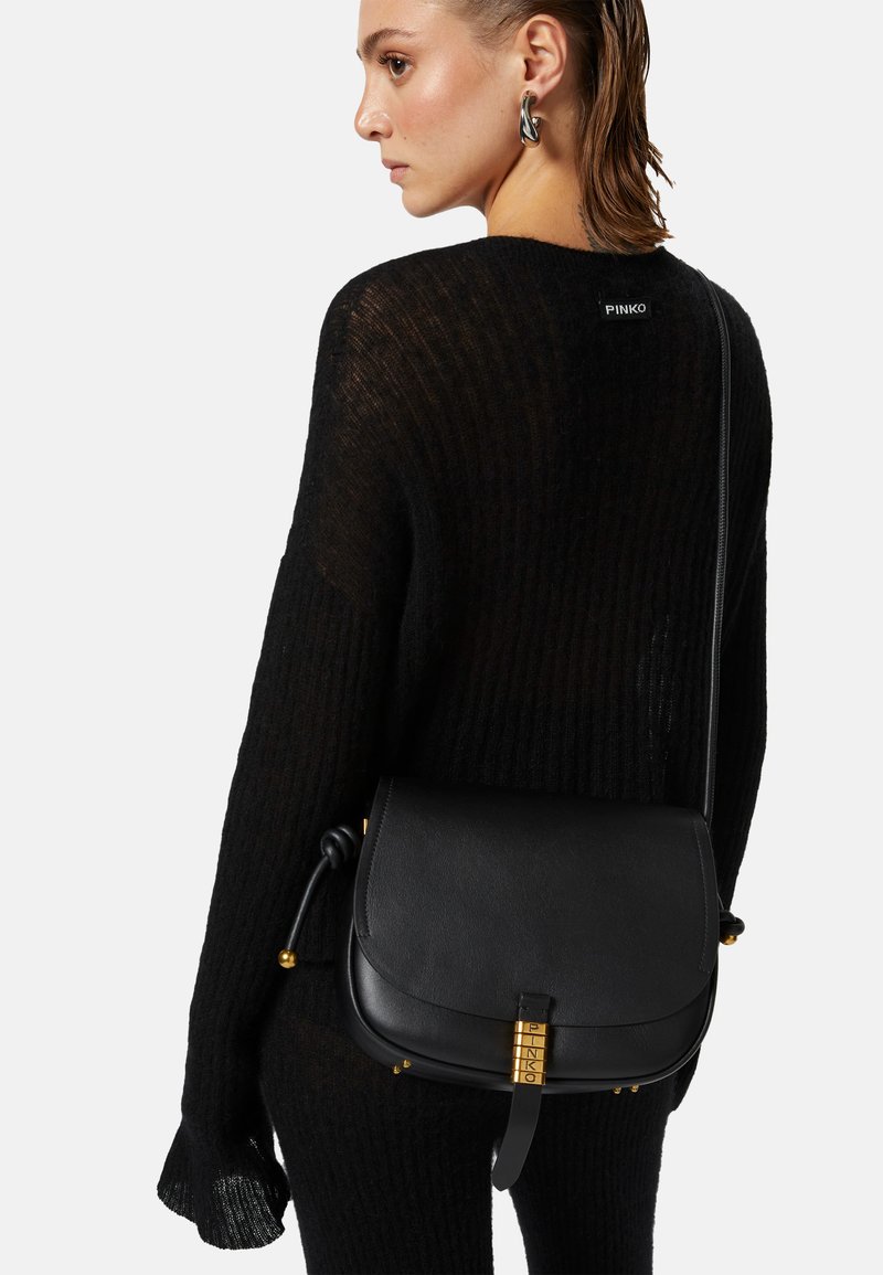 Borsa a tracolla nera in pelle con forma arrotondata, dotata di hardware in tonalità oro e una tracolla annodata, abbinata a un maglione in maglia nera con trama testurizzata.