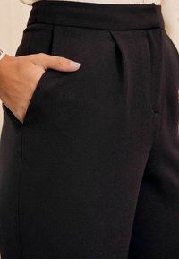 Zwarte broek met een gladde textuur en een licht taps toelopende pasvorm, voorzien van zijzakken en zichtbare voorplooien bij de taille.