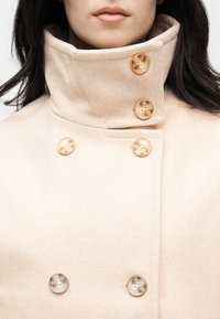 Γυναίκα που φοράει μπεζ μάλλινο παλτό με ψηλό γιακά και έξι μεγάλα στρογγυλά κουμπιά, κοντινή λήψη στο λαιμό και το άνω μέρος του στήθους.