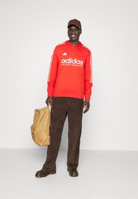 Felpa rossa Adidas con logo bianco, pantaloni in corduroy marroni, zaino marrone e scarpe marroni. Il modello indossa un cappello marrone abbinato.