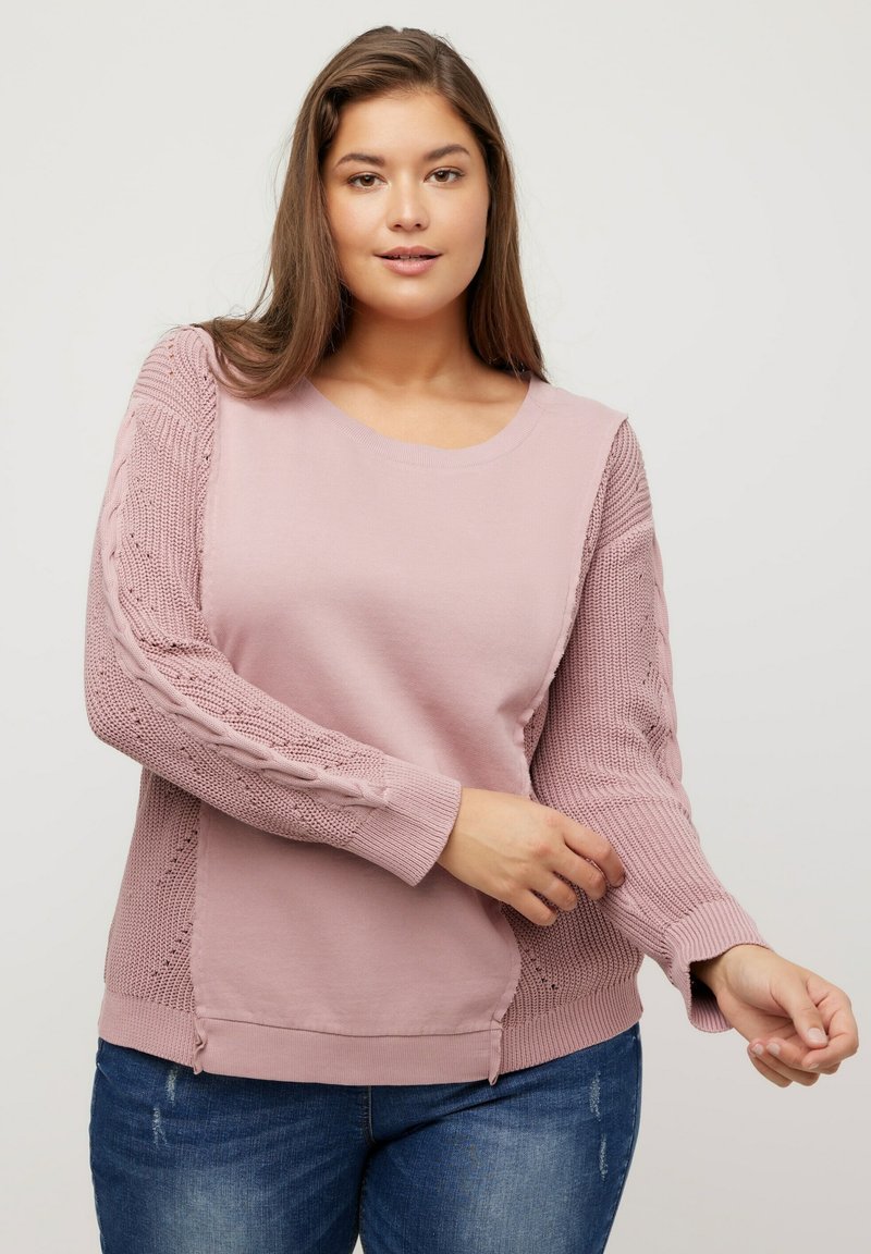 Vaaleanpunainen neuletakki pyöreällä kaula-aukolla, jossa on tekstuurilla koristellut köysineuleset hihat ja sileä vartalo. Sivuhalkiot lisäävät yksityiskohtia suunnitteluun.