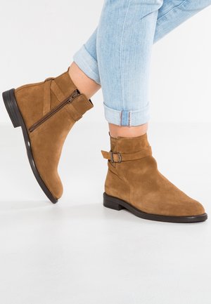 Botas de ante marrones hasta el tobillo con cierre de cremallera y correa ajustable. Suela plana y puntera redonda, colocadas sobre jeans celestes enrollados.