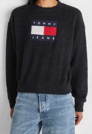 Personne portant un pull noir duveteux à manches longues avec un logo Tommy Jeans et un jean denim bleu clair.