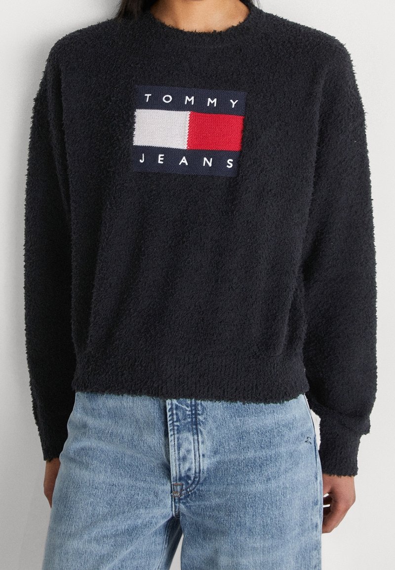 Personne portant un pull noir duveteux à manches longues avec un logo Tommy Jeans et un jean denim bleu clair.