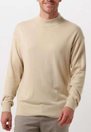 Homme souriant portant un pull beige à manches longues avec poignets et ourlet côtelés, associé à un pantalon gris à carreaux, debout sur fond blanc.