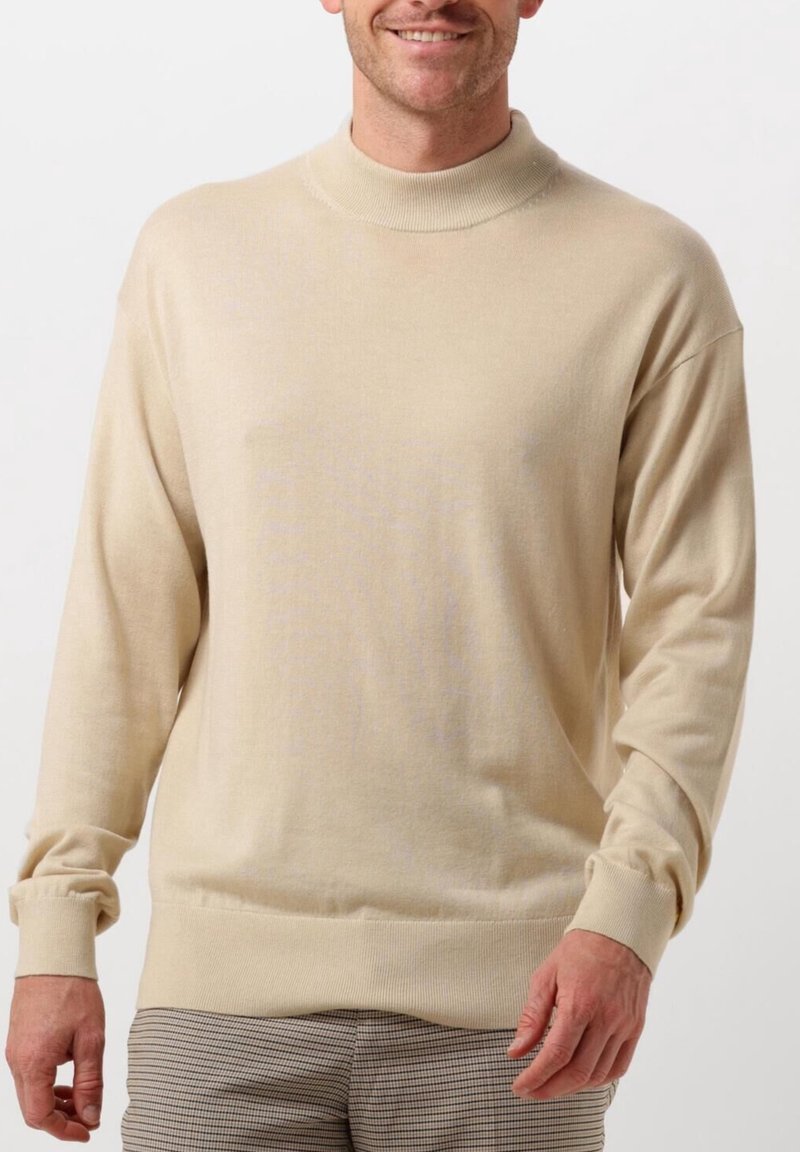Homme souriant portant un pull beige à manches longues avec poignets et ourlet côtelés, associé à un pantalon gris à carreaux, debout sur fond blanc.