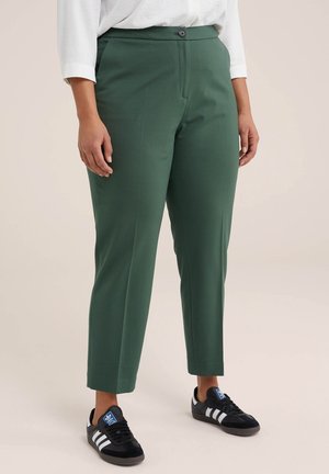 Woman wearing cropped green trousers, white long-sleeve shirt, and black Adidas sneakers with white stripes, standing on neutral background.