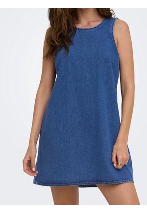 Donna con mini abito di jeans blu senza maniche e scollo rotondo, in piedi davanti a uno sfondo bianco neutro.