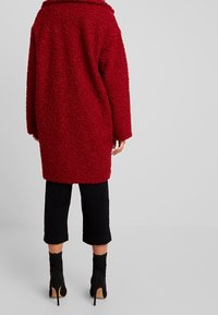 Červený chlupatý kabát volnějšího střihu s širokým límcem a dlouhými rukávy. Černé krátké kalhoty a vysoké kozačky celý vzhled doplňují.